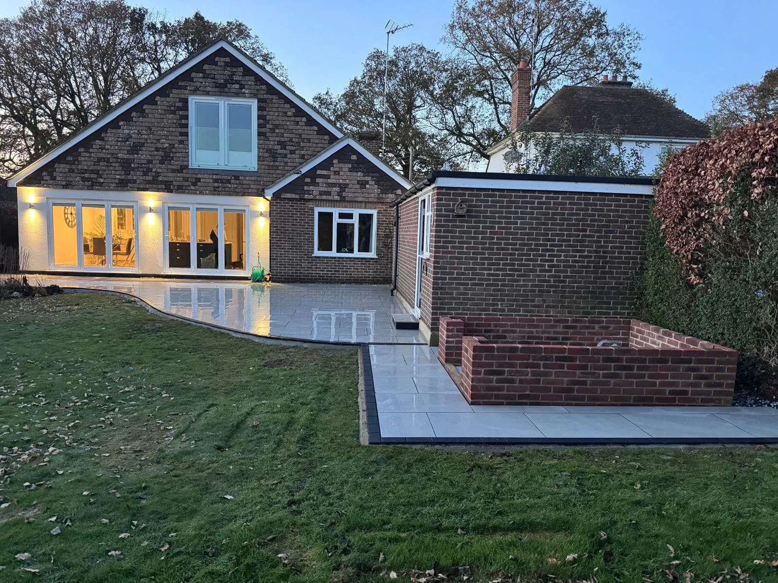 Back of a chalet bungalow at dusk with a new porcelain patio, brick raised bed and outdoor lights, finished by GPW